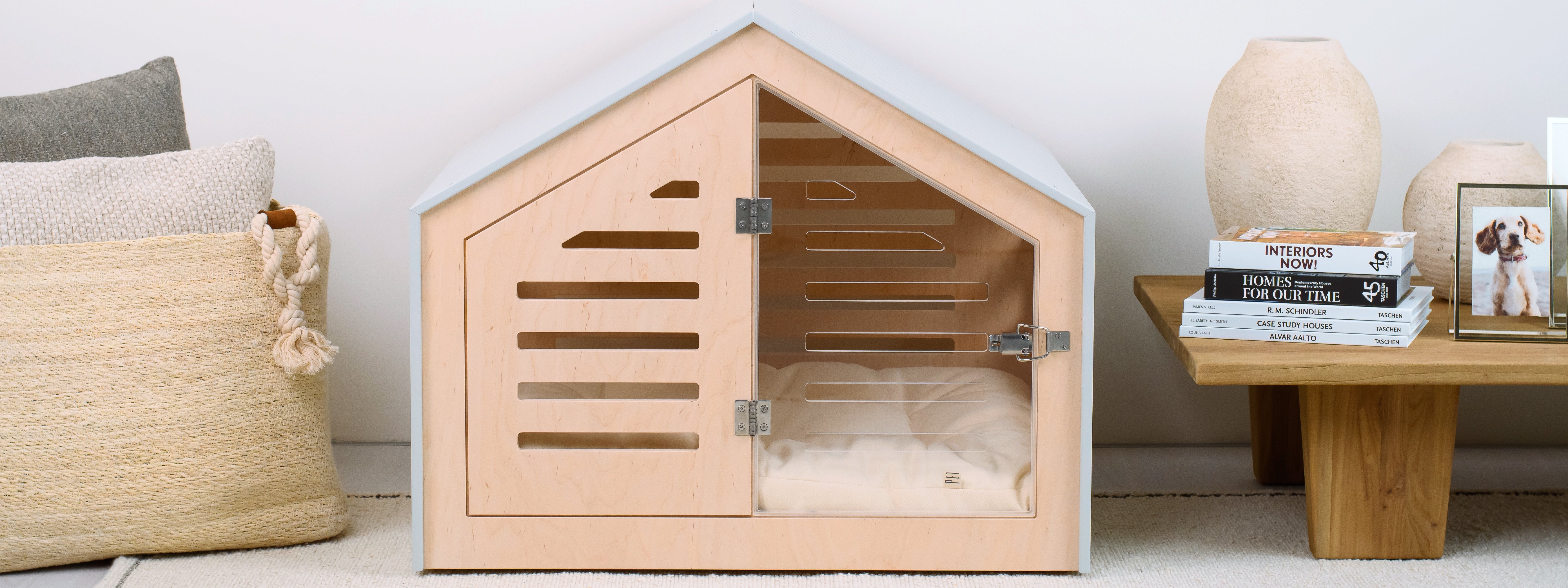 Wooden pet house with a bed inside, placed on a floor with a basket and books in the background.